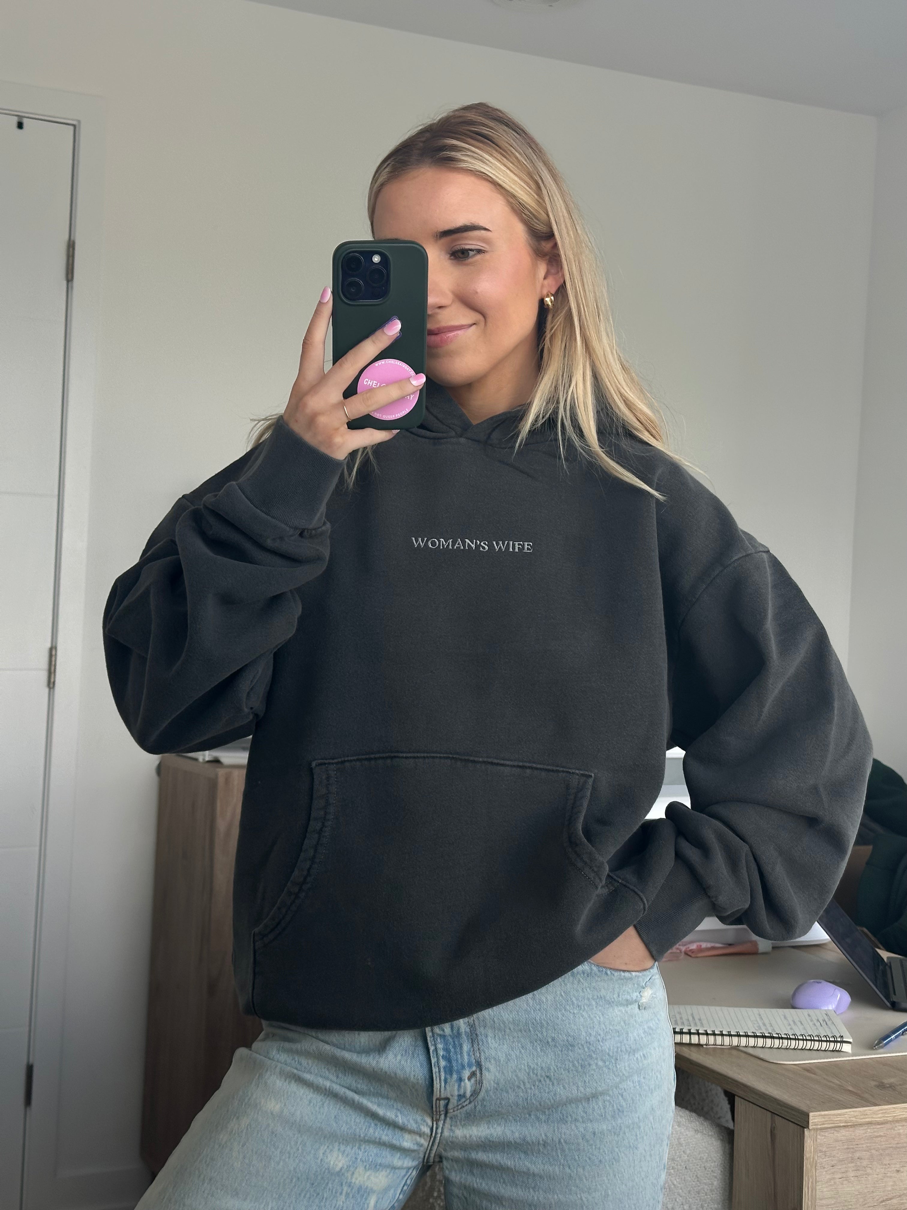 Person wearing a dark grey hoodie with 'WOMAN'S WIFE' text, standing in a room.