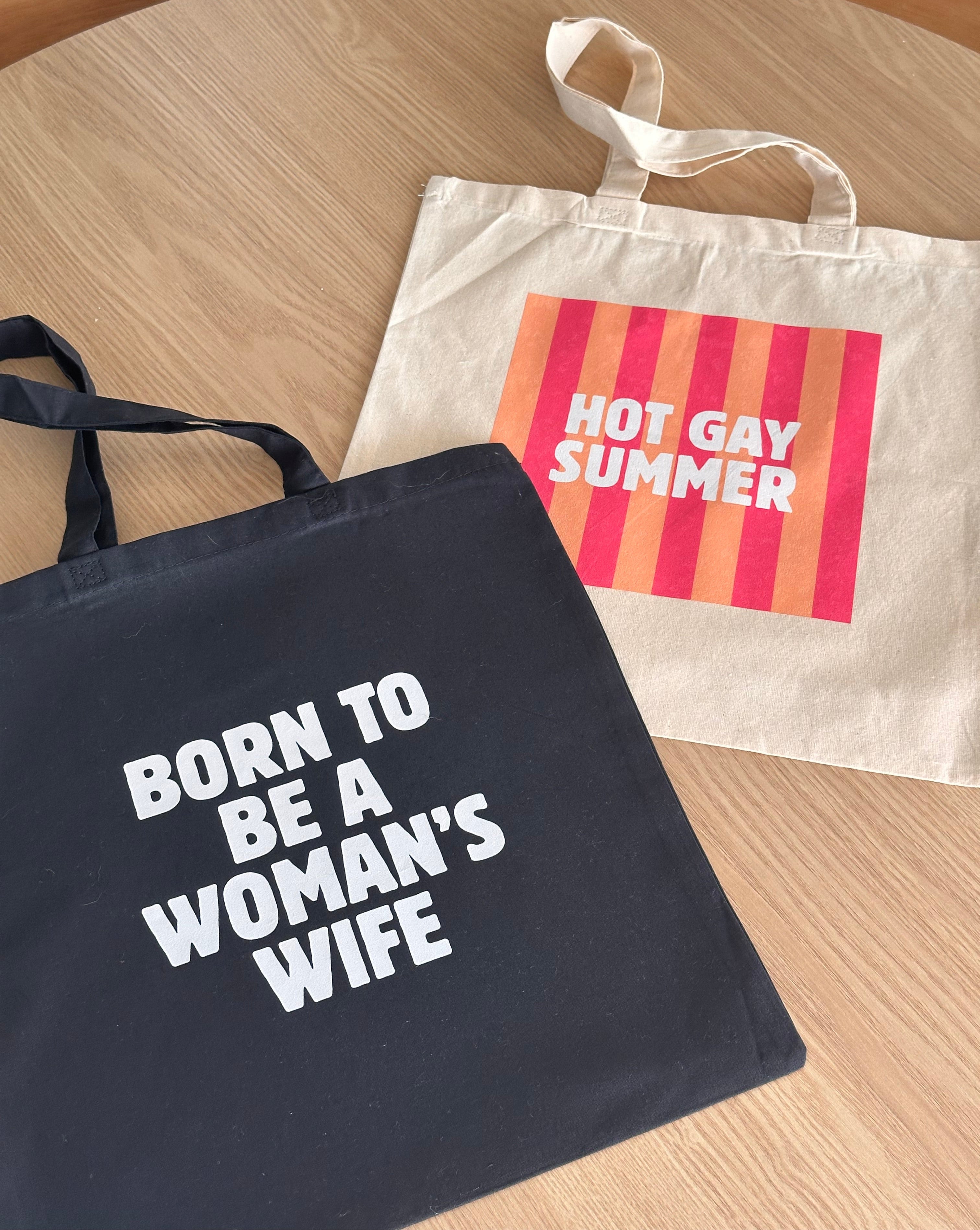 Black tote bag with 'Born to Be a Woman's Wife' text on a wooden surface, next to a beige tote bag with a colorful design and 'hot gay summer' text