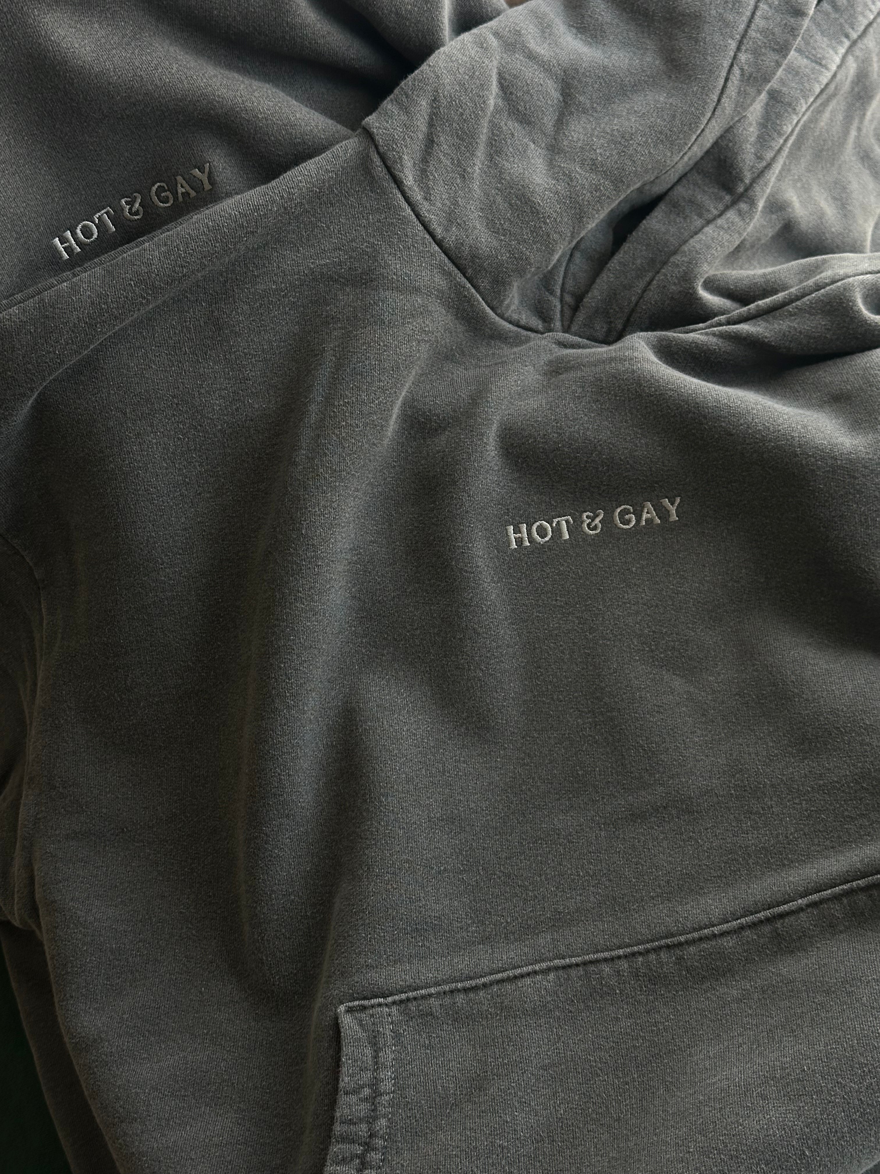 Dark grey hoodie with 'HOT & GAY' embroidery on a close-up of clothing.