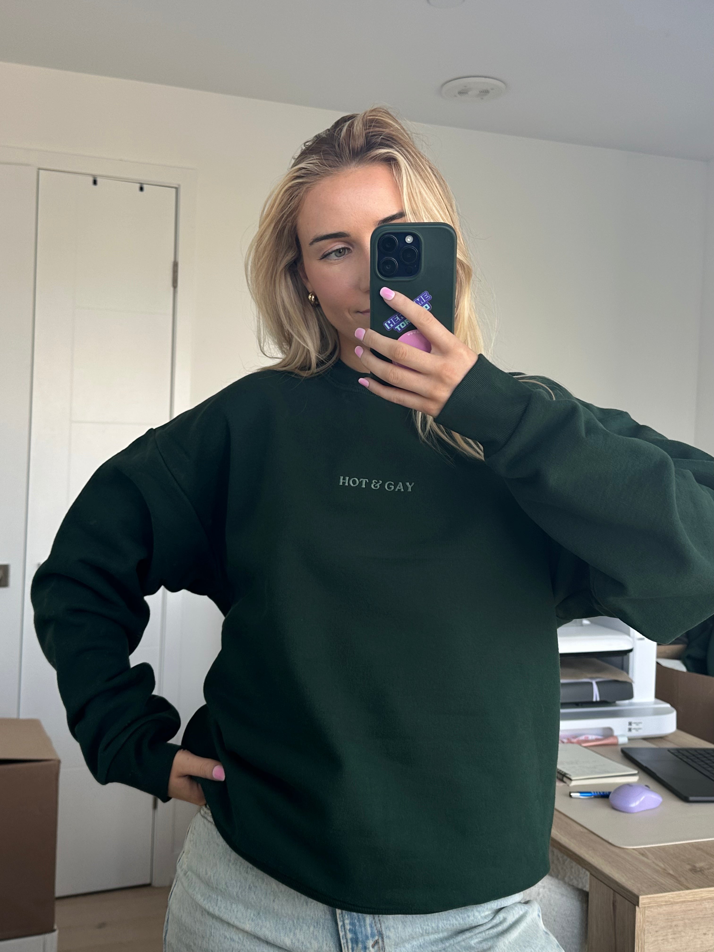 Person wearing a dark green sweatshirt with 'HOT & GAY' text, taking a mirror selfie.