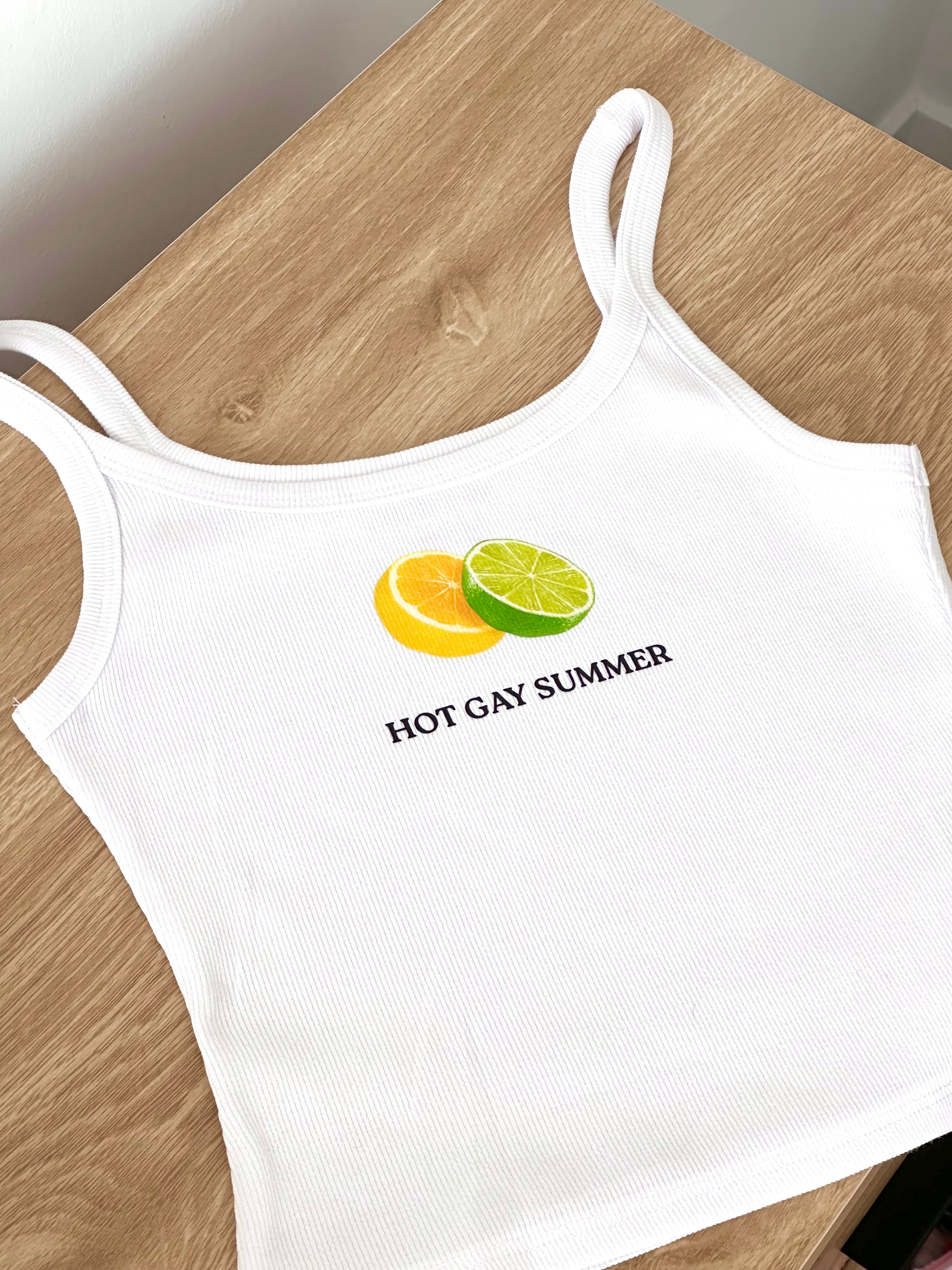 White tank top with fruit design and 'hot gay summer' text on a wooden surface