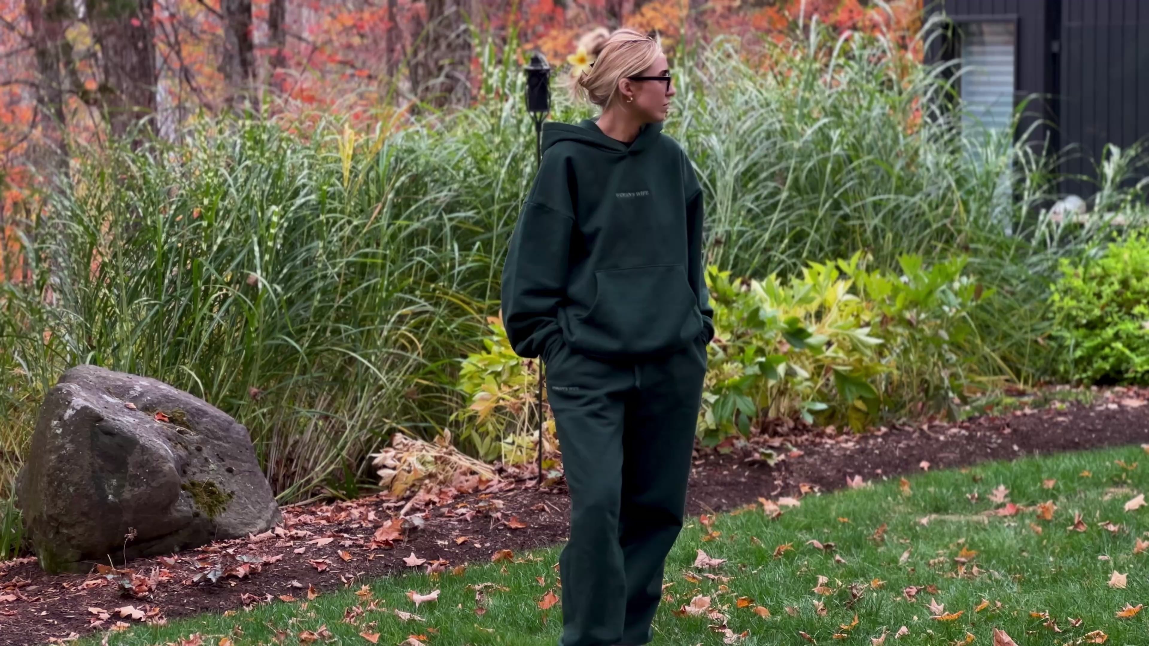 video of woman wearing 'woman's wife' dark green sweatsuit walking on grass with autumn greenery background