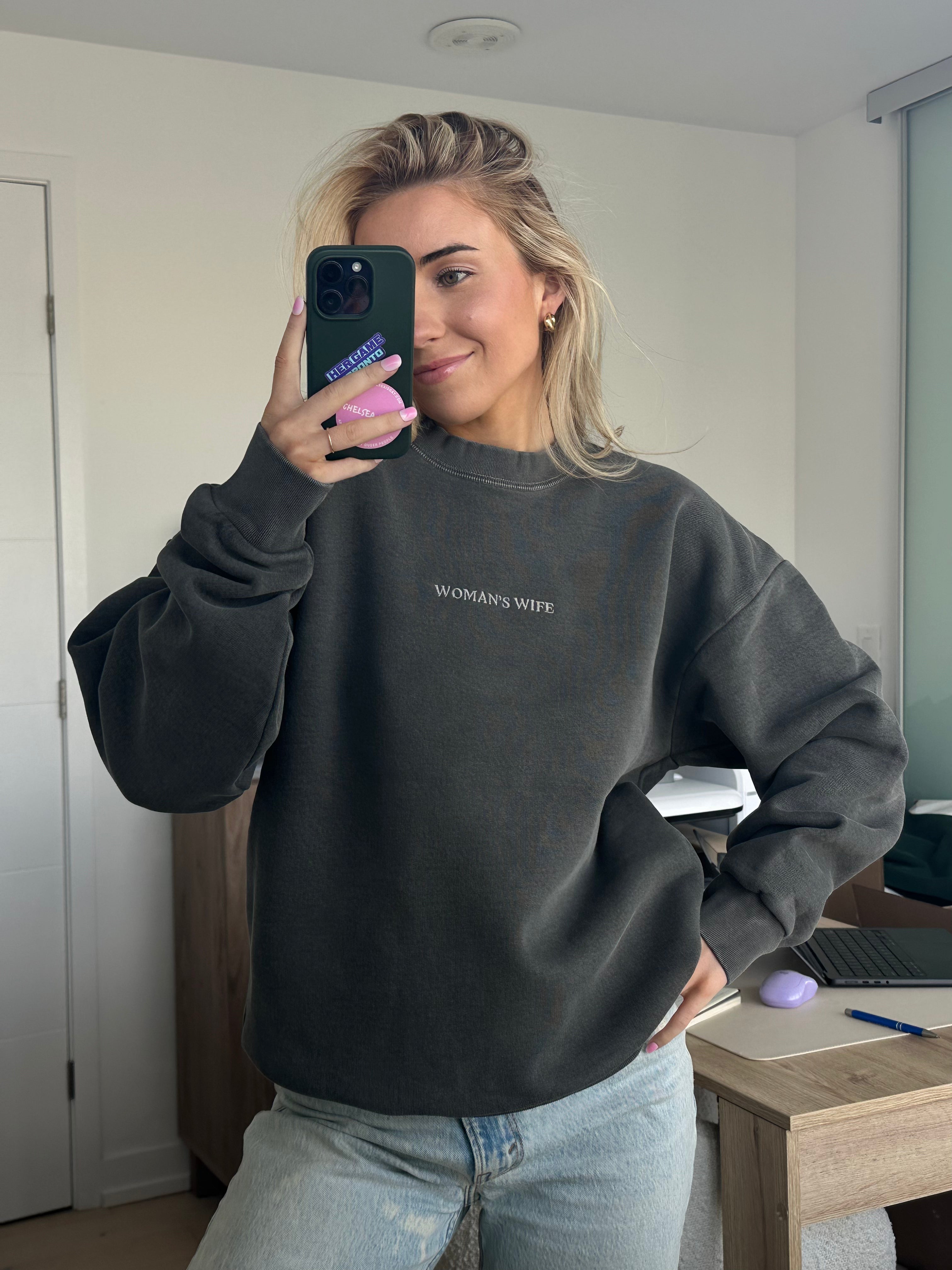 Person wearing a dark gray sweatshirt with 'WOMAN'S WIFE' text, holding a phone in a room.